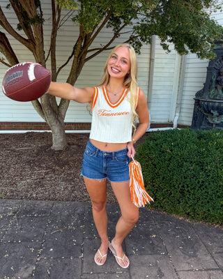 A smiling blonde woman stands outdoors on a paved walkway in front of a house, wearing a cream sleeveless crop top with orange trim and the word 'Tennessee' on the front, blue denim shorts, and flip-flops. She holds a brown football in one hand and an orange-and-white pom-pom in the other.