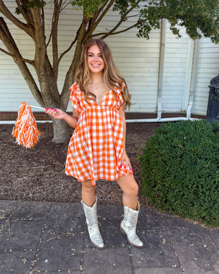 A woman stands outdoors in front of a white house and tree, wearing a bright orange and white gingham mini dress with flutter sleeves and holding an orange and white pom-pom. She is also wearing white cowboy boots.