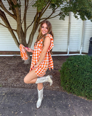 A woman posing outdoors in front of a white house wall and tree, wearing an orange and white gingham mini dress with ruffled straps and a matching headband, paired with white cowboy boots.