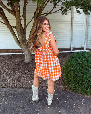 A woman wearing a short orange and white gingham dress with tie-shoulder straps and a back cutout, paired with white cowboy boots, standing outdoors on a paved walkway in front of a white house and a tree.