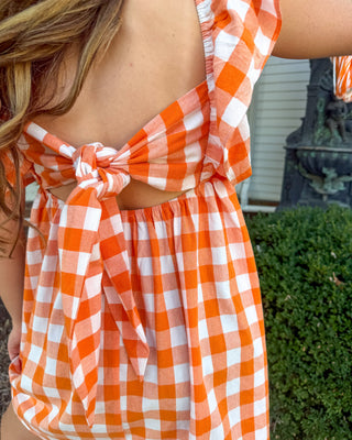 A close-up of a person wearing a bright orange and white gingham dress, showing the back tie detail and open back design in an outdoor setting.