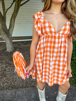 A woman wearing a bright orange and white gingham mini dress with ruffled sleeves, holding an orange and white pom-pom. She is standing outdoors in front of a house, wearing white cowboy boots.