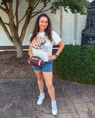 A woman stands outdoors on a paved walkway in front of a house, wearing a white graphic t-shirt with an orange and white football helmet design, blue denim shorts, and white sneakers, while holding a brown football.