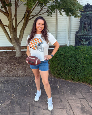 A woman stands outdoors on a paved walkway in front of a house, wearing a white graphic t-shirt with an orange and white football helmet design, blue denim shorts, and white sneakers, while holding a brown football.