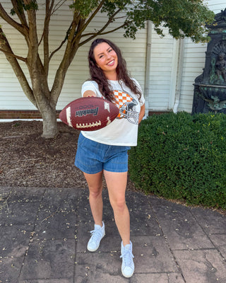 A woman stands outdoors on a paved walkway holding a brown football, wearing a white graphic t-shirt, denim shorts, and white sneakers. A tree, white house siding, and green shrubs are visible in the background.