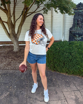A woman stands outdoors on a paved walkway holding a football, wearing a white graphic t-shirt with an orange and white checkerboard helmet design, blue denim shorts, and white sneakers.