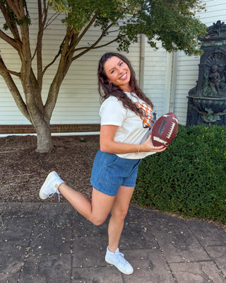 A woman outdoors on a paved walkway in front of a white building, holding a brown football. She is wearing a white t-shirt, denim shorts, and white sneakers, with a tree and greenery in the background.