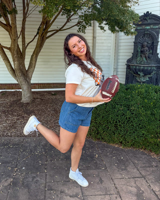 A woman outdoors on a paved walkway in front of a white building, holding a brown football. She is wearing a white t-shirt, denim shorts, and white sneakers, with a tree and greenery in the background.