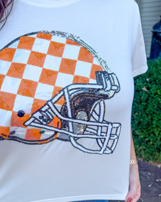 A white short-sleeve t-shirt featuring a large sequined football helmet graphic in orange, white, and black. The shirt is being worn by a person, with a blurred outdoor background of green foliage and a house.