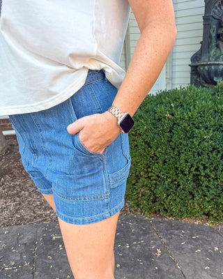 A person wearing light-wash blue denim shorts with a white sleeveless top and white sneakers, standing outdoors on a paved walkway with a green hedge and house siding in the background.