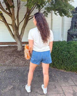 A woman stands outdoors on a paved walkway in front of a white house, viewed from behind. She is wearing a white t-shirt, blue denim shorts, and white sneakers, and is holding an orange and white pom-pom.