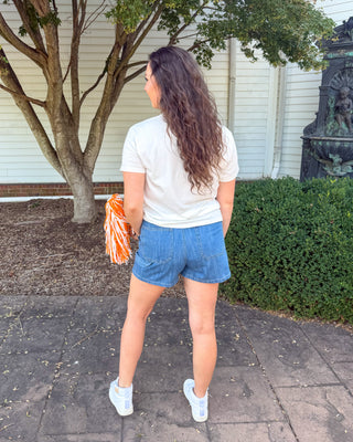 A woman stands outdoors on a paved walkway in front of a white house, viewed from behind. She is wearing a white t-shirt, blue denim shorts, and white sneakers, and is holding an orange and white pom-pom.