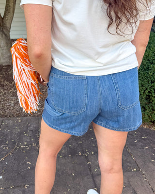 A person shown from behind wearing a white t-shirt and blue denim shorts, holding an orange and white pom-pom outdoors on a paved walkway near a tree.