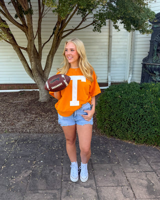 A young woman stands outdoors on a paved walkway in front of a house, holding a football. She is wearing a bright orange t-shirt with a large white letter 'T' on the front, light blue denim shorts, and white sneakers.