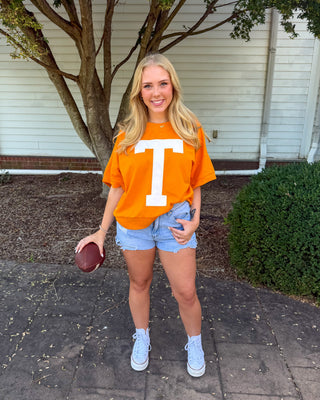 A young woman stands outdoors on a paved walkway in front of a house, wearing an oversized bright orange T-shirt with a large white letter 'T' on the front, light blue denim shorts, white sneakers, and holding a football.