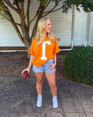 A woman stands outdoors on a paved walkway in front of a house, wearing an orange short-sleeve T-shirt with a large white letter 'T' on the front, denim shorts, white socks, and white sneakers, while holding a football.