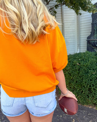 A person from behind wearing a bright orange oversized short-sleeve top and light blue denim shorts, holding a brown football outdoors next to a green hedge and white house siding.