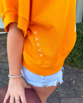A close-up lifestyle shot of a person wearing a bright orange sweatshirt with white stitched details resembling football stitching, paired with light blue denim shorts and holding a brown football.