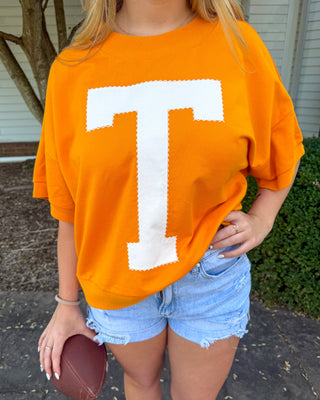 A woman wearing an oversized bright orange t-shirt with a large white block letter 'T' on the front, paired with light blue denim shorts and holding a brown football in an outdoor setting.