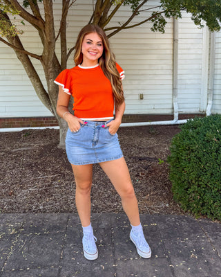 A young woman posing outdoors in front of a white building and tree, wearing a bright orange short-sleeve ringer t-shirt with white trim, a light wash denim mini skirt, and light blue sneakers.