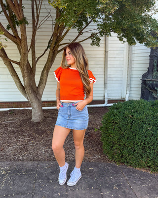 A woman posing outdoors in a bright orange short-sleeve crop top with white ruffle details on the sleeves, paired with a denim mini skirt and white sneakers.