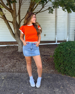 A woman stands outdoors in front of a white house wall and tree, wearing a bright orange short-sleeve t-shirt with white ruffle trim, a light blue denim mini skirt, white socks, and white sneakers.