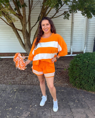 A woman stands outdoors on a paved walkway in front of a house, wearing an orange and white athletic-style lounge set with a matching pom-pom. The outfit consists of a loose short-sleeve top with wide sleeves and matching shorts with white stripes.