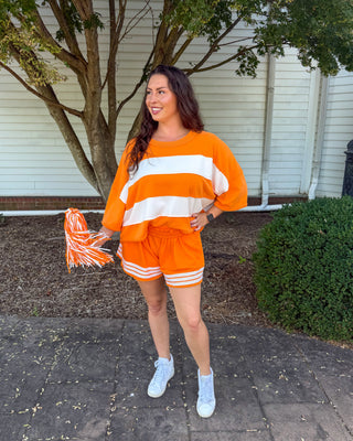 A woman stands outdoors on a paved walkway in front of a house, wearing an orange and white striped long-sleeve top and matching orange shorts with white stripes, holding an orange and white pom-pom.