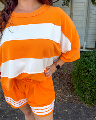 A person wearing a bright orange and white striped lounge set, featuring an oversized short-sleeve top and matching shorts with white stripes near the hem, photographed outdoors on a patio with shrubs in the background.