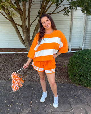 A woman stands outdoors on a paved walkway in front of a house, wearing an orange and white striped long-sleeve top and matching orange shorts with white stripes. She is holding an orange and white pom-pom.