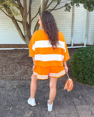 A woman stands outdoors on a paved walkway in front of a white building and tree, wearing an orange and white striped short-sleeve top and matching orange shorts with white stripes. She is seen from the back and holds an orange and white pom-pom.