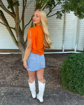 A woman stands outdoors in front of a white house wall and tree, wearing a bright orange short-sleeve top with fringe, a light blue denim mini skirt, and white cowboy boots.