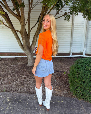 A woman standing outdoors in front of a white house wall and tree, wearing a bright orange short-sleeve fringed top, a light blue denim mini skirt, and white cowboy boots.
