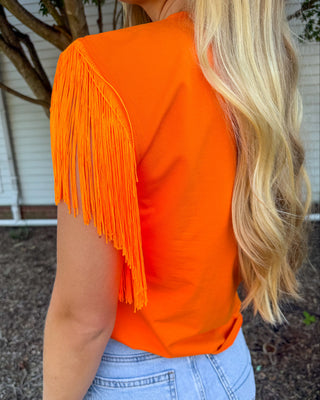 A woman wearing a bright orange short-sleeve top with fringe detailing along the shoulder, paired with light blue denim shorts, photographed outdoors in front of a house.