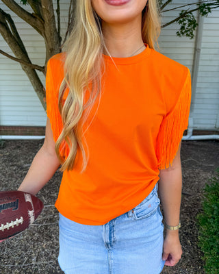 A woman wearing a bright orange short-sleeve t-shirt with fringe details on the sleeves, paired with a light blue denim mini skirt. She is holding a football in a backyard setting with a house and tree in the background.