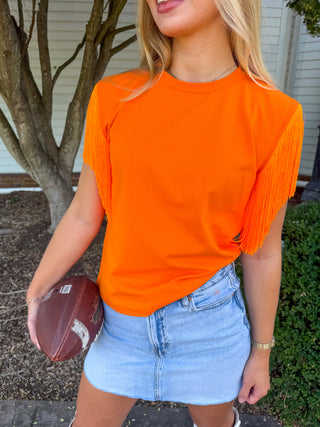 A woman wearing a bright orange short-sleeve t-shirt with fringe details on the sleeves, paired with a light blue denim mini skirt, holding a football in an outdoor setting.