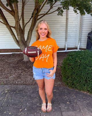 A woman stands outdoors holding a football, wearing a bright orange graphic t-shirt with the words 'GAME DAY' in white script, paired with denim shorts and sandals.