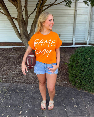 A woman stands outdoors on a paved walkway in front of a house, wearing a bright orange graphic T-shirt that reads 'GAME DAY' in white script, paired with denim shorts and sandals. She is holding a football.