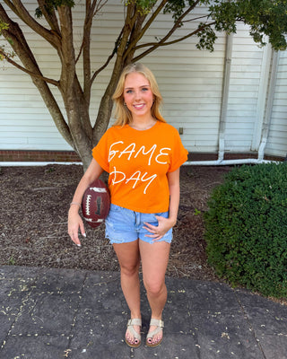 A woman stands outdoors in front of a house, wearing a bright orange t-shirt with 'GAME DAY' printed in white script across the chest, paired with denim shorts and sandals, while holding a football.