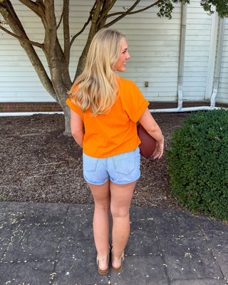 A woman stands outdoors on a paved walkway in front of a house, wearing a bright orange short-sleeve top and light blue denim shorts, holding a football.