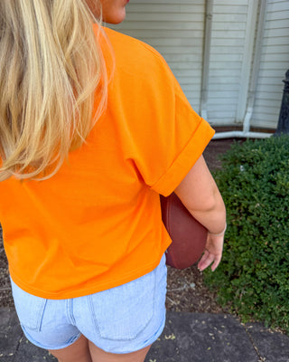 A blonde girl seen from behind wearing a bright orange short-sleeve t-shirt and light blue denim shorts, holding a brown football outdoors next to a green bush.