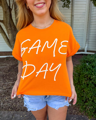 A woman wearing a bright orange short-sleeve t-shirt with 'GAME DAY' printed in white script across the front, paired with light blue denim shorts, standing outdoors in front of a house and trees.