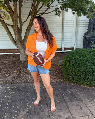 A woman stands outdoors on a paved walkway holding a football, wearing an orange zip-up jacket over a white tank top, denim shorts, and white sandals. Trees, a white house wall, and a dark decorative fountain are visible in the background.