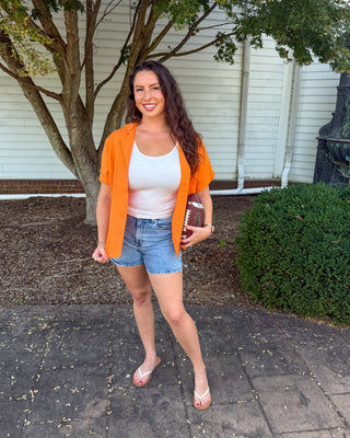 A woman stands outdoors on a paved walkway in front of a white house, wearing a bright orange short-sleeve button-up shirt over a white tank top, denim shorts, and pink flip-flop sandals, while holding a football.