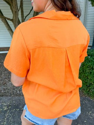 A person viewed from the back wearing a bright orange short-sleeve collared shirt and denim shorts outdoors in front of a house and tree.