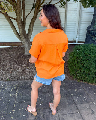 A woman wearing a bright orange short-sleeve button-up shirt and denim shorts, standing outdoors on a paved walkway in front of a house and a tree.