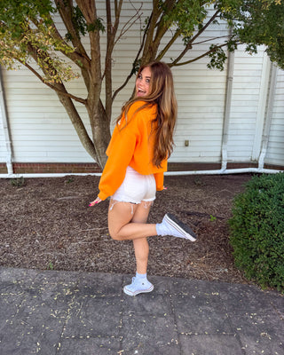 A woman posing outdoors in a bright orange cropped hoodie, white shorts, and white sneakers, standing on a paved walkway in front of a white house and tree.