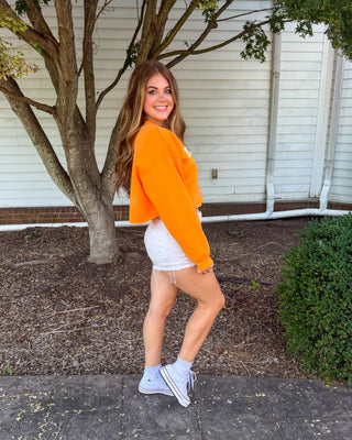 A woman posing outdoors on a paved walkway in front of a white house, wearing a bright orange cropped long-sleeve top, white shorts, and white sneakers.
