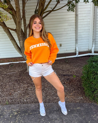 A woman posing outdoors in front of a house, wearing an orange crewneck sweatshirt with 'TENNESSEE' printed in white across the chest, white denim shorts, and white sneakers.