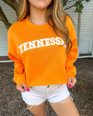 A woman wearing a bright orange crewneck sweatshirt with 'TENNESSEE' printed in white arched block letters across the chest, paired with white denim shorts.